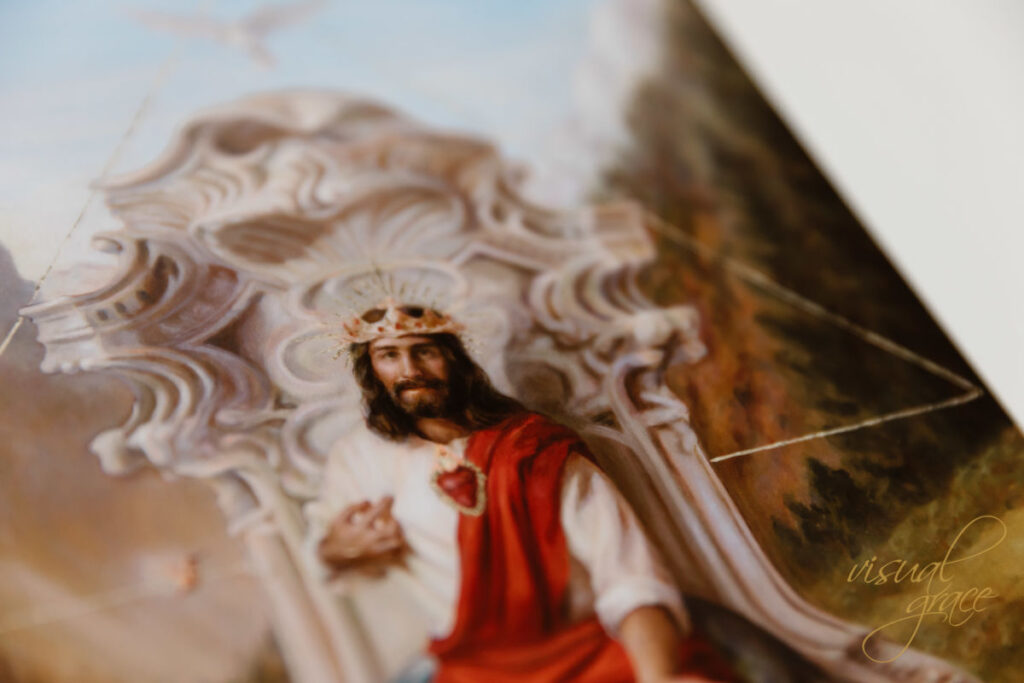 Jesus sitting one a throne enrobed in a white tunic and a red sash flowing from his shoulder to his feet. He is pointing to his Sacred Heart with one hand and reaching out to the viewer with the other hand.